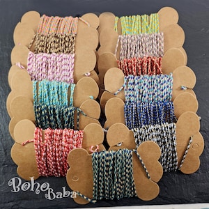 Puede incluir: Una colección de 12 carretes de hilo de bordar de colores, cada uno enrollado en un carrete de cartón marrón con un recorte en forma de corazón. El hilo es una mezcla de colores, incluyendo rojo, azul, verde, amarillo y blanco.