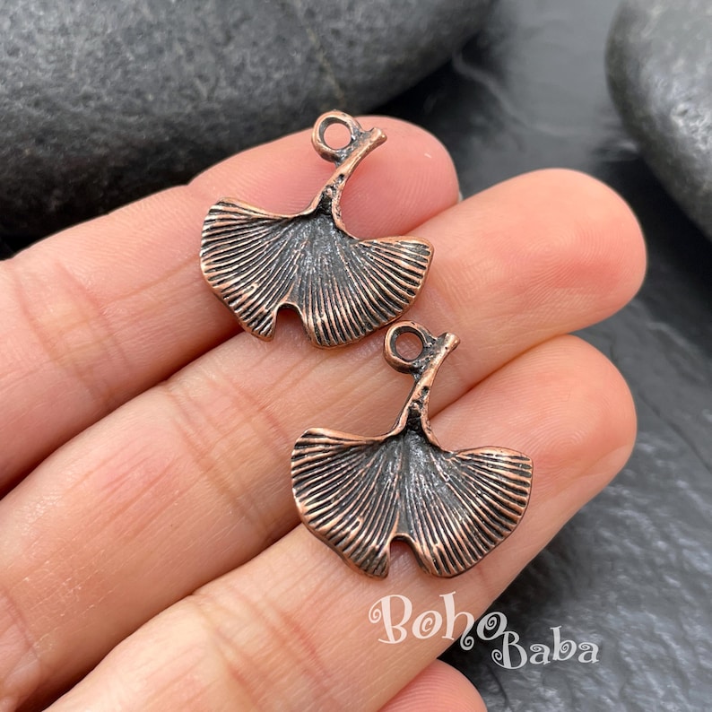 A close-up photograph displays two antique copper plated ginkgo leaf charms resting on a person's fingers against a dark rock background.