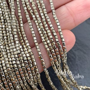 Può includere: Primo piano di fili di perline quadrate dorate metalliche. Le perline sono infilate insieme, creando una superficie strutturata e riflettente. Le perline sono piccole e di forma uniforme, ideali per la creazione di gioielli o progetti artigianali.