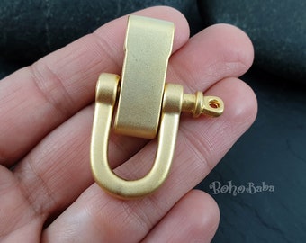 May include: A gold-colored metal shackle with a U-shaped base and a rectangular top. The shackle has a hinged design and a small, cylindrical pin on one end. The metal has a matte finish.