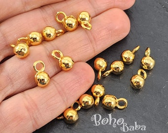 May include: A collection of small, shiny gold-colored jewelry findings. Each finding has a rounded bead shape with a small loop attached. The findings are scattered across a surface, with some held in a hand. The text "Boho Baba" is visible.