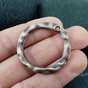 Pode incluir: Um anel de cobre martelado com um furo para prender um pingente ou um amuleto. O anel tem um diâmetro de aproximadamente 2,5 cm.