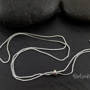 Könnte beinhalten: Eine silberne Kettenkette mit einem Schiebeverschluss. Die Kette besteht aus kleinen, miteinander verbundenen Kreisen. Die Kette liegt auf einer dunkelgrauen Oberfläche.