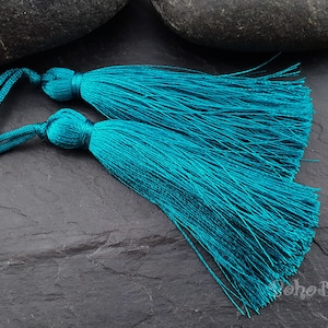 Peut inclure: Deux glands de couleur bleu sarcelle avec de longues franges. Les glands sont liés ensemble et reposent sur une surface gris foncé.