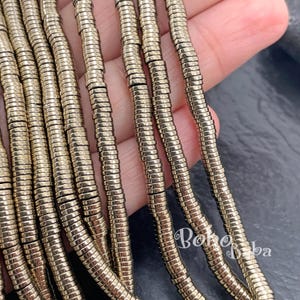 May include: Close-up of several strands of gold-colored, segmented beads. The beads are cylindrical and have a metallic sheen. The beads are held in a hand, with a dark background. The beads are likely for jewelry making.
