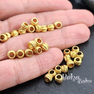 May include: A close-up of a hand holding a large quantity of gold-colored metal beads. The beads are round and have a textured surface. The beads are approximately 1/2 inch in diameter.