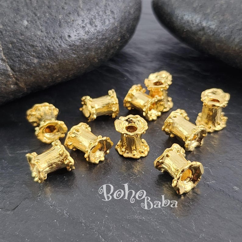 May include: A group of 12 gold-colored metal bead caps with a decorative, scalloped edge. The bead caps are arranged on a dark gray surface.