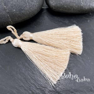 May include: Two cream-coloured silk tassels with looped cords. The tassels are made of many strands of silky material, and the top is a rounded, wrapped design. The tassels are on a dark, textured surface.