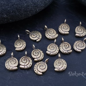 Puede incluir: Una colección de 15 abalorios de metal dorado con un diseño en espiral. Los abalorios están esparcidos sobre una superficie gris oscura.