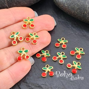 May include: Small gold-coloured cherry charms with red and green enamel detailing. These charms, ideal for jewellery making, feature a pair of red cherries and green leaves. The charms are displayed on a dark surface.