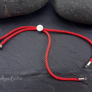May include: Red adjustable cord bracelet with silver metal ends and a silver metal slider. The bracelet is on a dark gray surface.