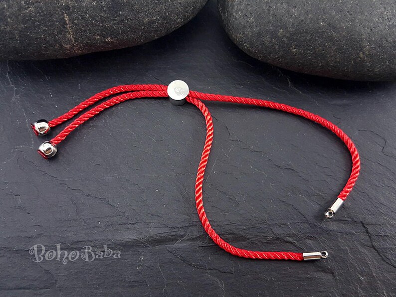 May include: Red adjustable cord bracelet with silver metal ends and a silver metal slider. The bracelet is on a dark gray surface.