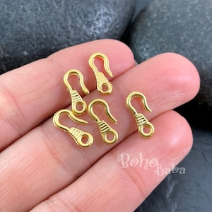 Könnte beinhalten: Sechs goldfarbene Metall-Hummerverschlüsse, jeweils mit einer strukturierten Oberfläche, werden auf einer Hand gezeigt. Die Verschlüsse sind etwa 2,5 cm lang.