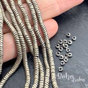 Puede incluir: Primer plano de hebras de cadena plateadas en forma de serpiente y pequeñas cuentas redondas. La cadena tiene un diseño ondulado. Las cuentas están dispersas sobre una superficie oscura. El texto "BohoBaba" es visible en la esquina inferior derecha.