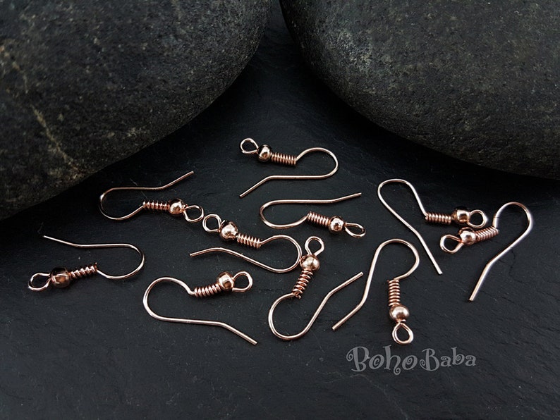 May include: A close-up of rose gold earring hooks. The hooks are made of metal and have a spring-loaded closure. The hooks are arranged on a black surface.