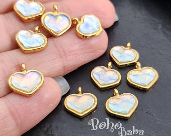 Puede incluir: Surtido de diez dijes de corazón dorados con inserciones de vidrio blanco y azul iridiscente. Los dijes se muestran en una mano.