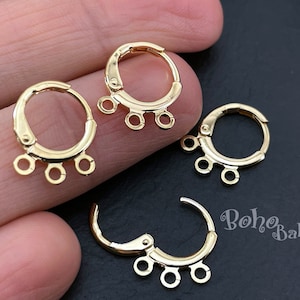 Puede incluir: Pendientes de aro dorados con tres pequeños bucles para sujetar abalorios o colgantes. Los pendientes se muestran en primer plano, con varios pares visibles. Tienen un acabado brillante y un diseño sencillo.