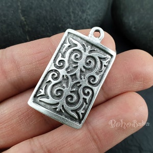 Puede incluir: Pendiente de plata con un diseño intrincado y ornamentado. El pendiente es de forma rectangular y tiene un bucle en la parte superior para sujetarlo a una cadena.