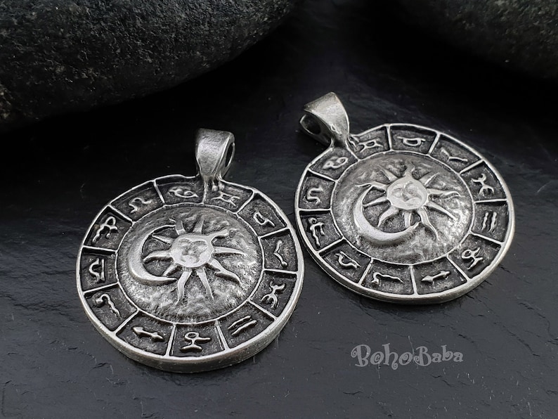 May include: Two silver pendants with a sun and moon design surrounded by a circle of zodiac symbols. The pendants are on a dark gray background.