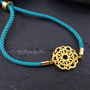May include: Teal blue cord bracelet with a gold-toned filigree charm. The charm is a circular design with a center hole.