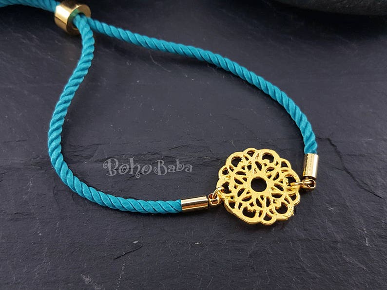 May include: Teal blue cord bracelet with a gold-toned filigree charm. The charm is a circular design with a center hole.