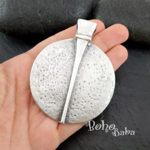 May include: A silver-toned pendant with a textured, circular design and a vertical bar accent. The pendant has a loop at the top for a chain. The BohoBaba logo is visible in the bottom right corner.