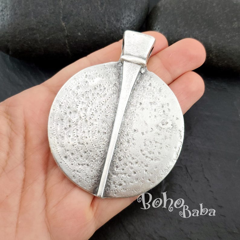 May include: A silver-toned pendant with a textured, circular design and a vertical bar accent. The pendant has a loop at the top for a chain. The BohoBaba logo is visible in the bottom right corner.