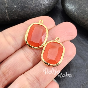 May include: Two orange-red gemstone pendants with gold-colored, textured bezels. Each pendant has a rectangular shape with rounded corners and a small loop at the top. The pendants are displayed on a light-colored background.
