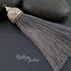 Puede incluir: Un borla gris con una parte superior de metal plateado con un diseño intrincado. La borla está sobre un fondo gris oscuro.