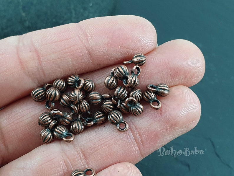 May include: A pile of small, copper-colored metal beads with a decorative, ribbed design. Each bead has a small loop at the top for attaching to jewelry or other crafts.