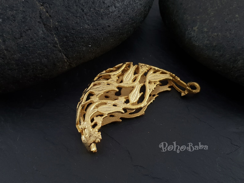 May include: Gold-toned metal pendant with a delicate, intricate design resembling a stylized floral pattern. The pendant has a loop at the top for attaching to a chain or cord.