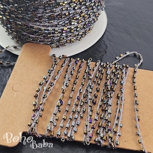 Puede incluir: Un carrete de hilo gris con cuentas doradas y plateadas iridiscentes. El hilo está enrollado alrededor de un carrete de cartón y las cuentas están enhebradas en el hilo. Las cuentas son pequeñas y redondas, y tienen un brillo metálico. El hilo mide aproximadamente 3 metros.