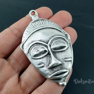 May include: Silver pendant featuring a detailed African mask design. The mask has a closed-eye expression and a textured surface.