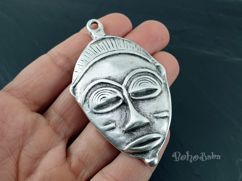May include: Silver pendant featuring a detailed African mask design. The mask has a closed-eye expression and a textured surface.