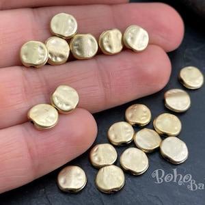 May include: A collection of gold-toned metal beads with a hammered finish. The beads are round and have a hole in the centre for stringing. The beads are scattered on a hand and a dark surface.