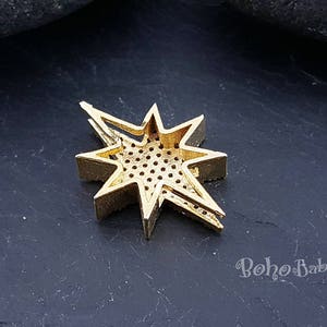 Puede incluir: Pendiente de metal dorado en forma de estrella con un dise&ntilde;o geom&eacute;trico y un centro estampado. El pendiente est&aacute; sobre una superficie gris oscura.