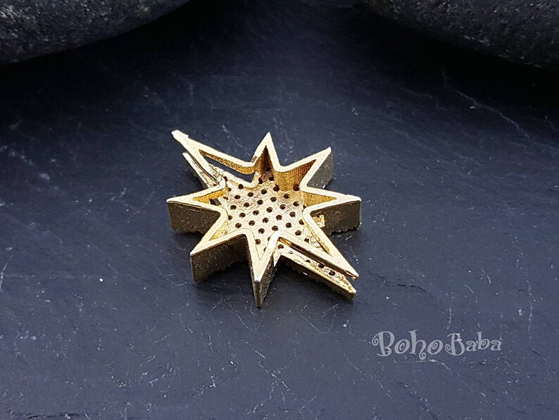 Puede incluir: Pendiente de metal dorado en forma de estrella con un dise&ntilde;o geom&eacute;trico y un centro estampado. El pendiente est&aacute; sobre una superficie gris oscura.