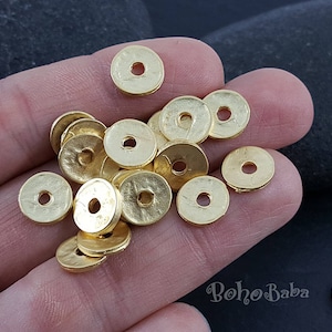 Könnte beinhalten: Ein Haufen goldfarbener Metallperlen, jede mit einem runden Loch in der Mitte. Die Perlen sind in einem zufälligen Muster auf einer Hand angeordnet.