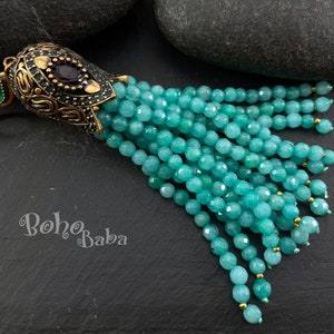 Könnte beinhalten: Eine türkisfarbene Edelstein-Quastenkette mit einem verzierten goldfarbenen Anhänger mit einem roten Edelstein. Der Anhänger hat eine Schlaufe zum Anbringen an einer Kette oder Schnur. Die Quaste besteht aus vielen kleinen, facettierten Türkisperlen.