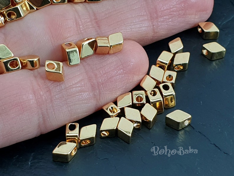 May include: A close-up of a pile of gold-colored metal beads. The beads are square and have a hole in the center. The beads are on a hand and a dark surface. The text "Boho Baba" is visible in the lower right corner of the image.