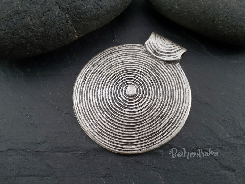 May include: Silver pendant with a circular design featuring concentric circles. The pendant has a loop at the top for attaching to a chain.