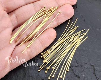 May include: A close-up of a large quantity of gold-colored head pins with round heads. The pins are arranged in two groups, one on top of the other.