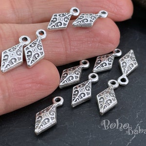 May include: A close-up of ten silver diamond-shaped charms with intricate designs. The charms have a loop at the top for attaching to jewellery or other crafts.