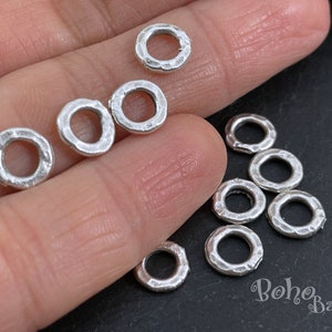 Puede incluir: Primer plano de una colección de anillos de salto de plata. Los anillos son todos del mismo tamaño y tienen una textura martillada. Los anillos están dispuestos en círculo sobre una superficie negra.