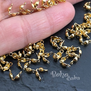 Op de afbeelding: Een close-up van een stapel gouden metalen sluitingen. De sluitingen zijn klein en hebben een afgeronde vorm. Ze liggen verspreid op een zwart oppervlak.