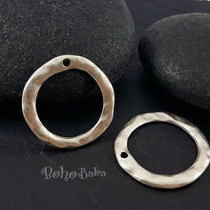 以下が含まれることがあります： ハンマーで叩いたような質感の銀色の金属製のリングが2つ。リングは丸く、上部にジュエリーを取り付けるための穴が開いています。