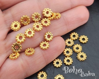 May include: A close-up of a hand holding a large quantity of gold-colored metal spacer beads. The beads are round with a small hole in the center and have a decorative design of small raised dots around the edge.