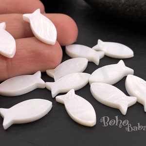Può includere: Una collezione di ciondoli a forma di pesce in madreperla bianca. I ciondoli sono lunghi circa 2,5 cm e hanno una finitura liscia e lucida.