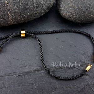 以下が含まれることがあります： 黒色のコードブレスレットに、金色の金属製のアクセントと小さな丸いビーズが2つ付いています。ブレスレットは、スライド式の結び目で調節できます。