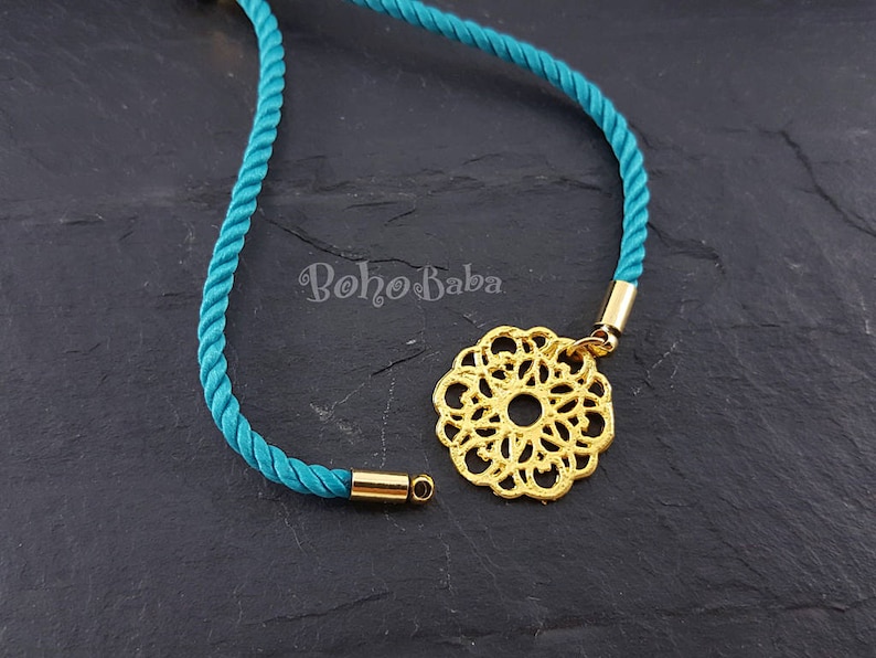 May include: A turquoise cord necklace with a gold filigree pendant. The pendant is a circular design with a small hole in the center.
